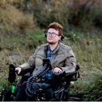 Jamie sits in their electric wheelchair. Behind them are bushes. They are wearing glasses and have dark red hair, and a red beard. They are dressed in a leather jacket, and look steadily at the camera
